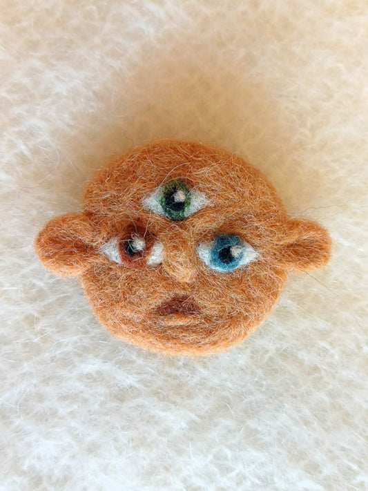 Small needle felted face with dark beige skin and 3 eyes; one green, one brown and one blue. His ears stick out either side and he carries a surprised expression.