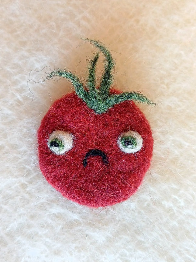 Needle felted wool red tomato brooch with a sad face on a white knitted fabric background. The brooch has large bulging eyes, a C shaped frown, and a tuft of fluffy green leaf 'hair'