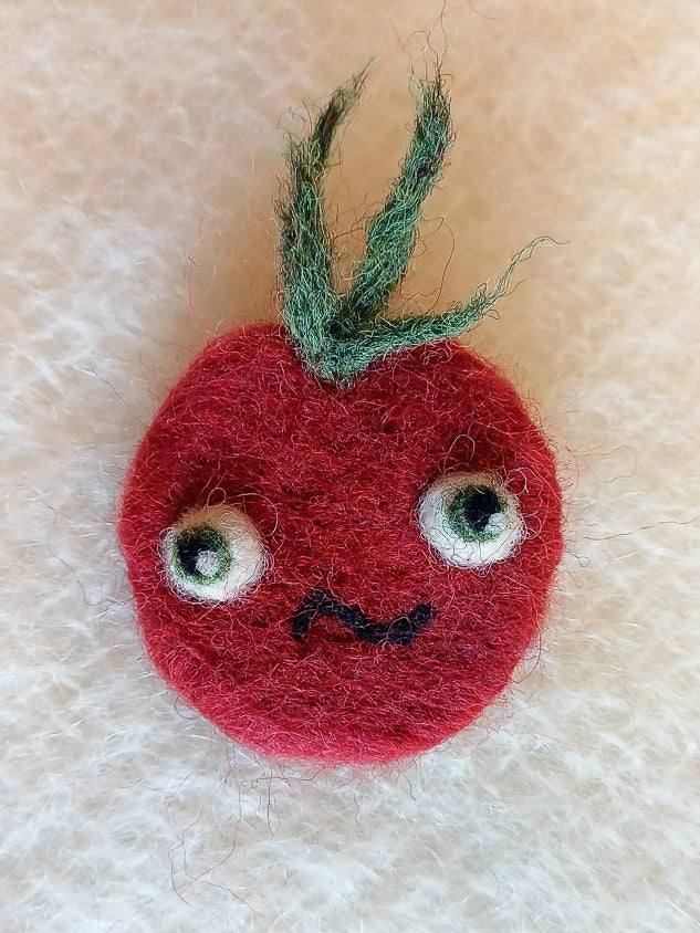 Needle felted wool red tomato brooch with a confused face on a white knitted fabric background. The brooch has large bulging eyes, a S shaped mouth, and a tuft of fluffy green leaf 'hair'
