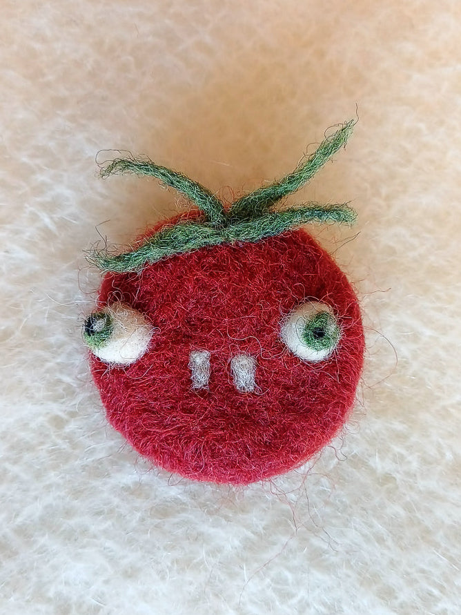 Needle felted wool red tomato brooch with buckteeth on a white knitted fabric background. The brooch has large bulging eyes, two white teeth, and a tuft of fluffy green leaf 'hair'