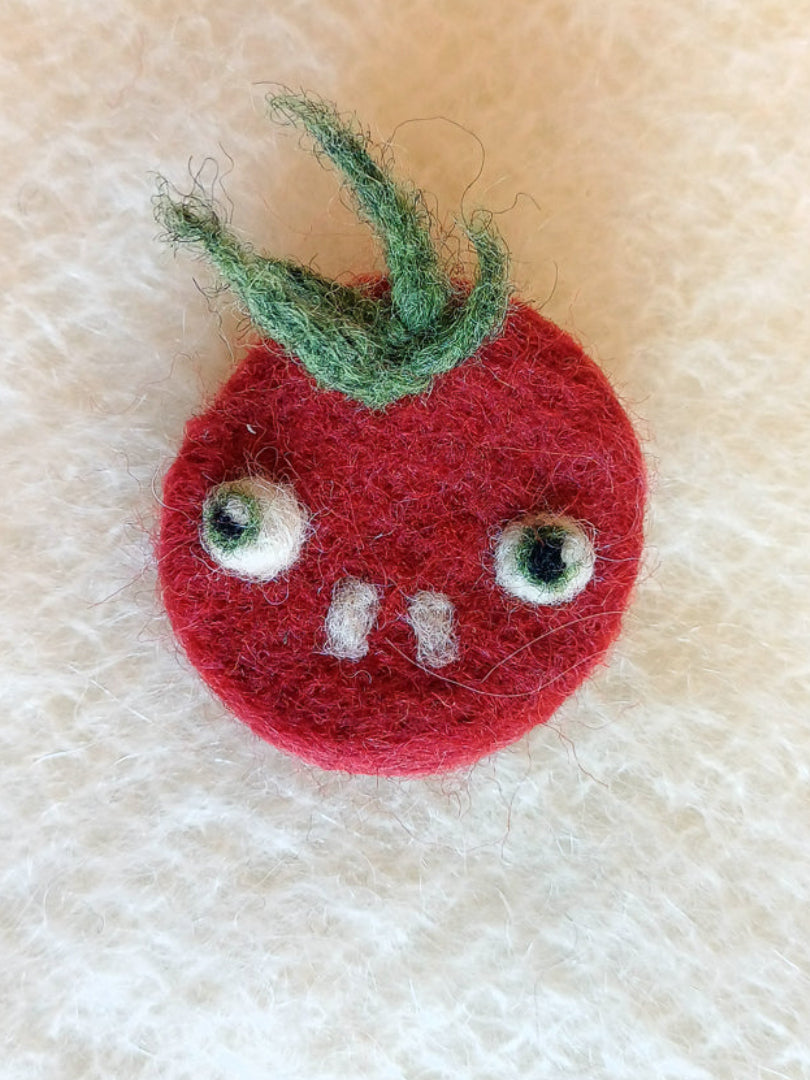 Needle felted wool red tomato brooch with buckteeth on a white knitted fabric background. The brooch has large bulging eyes, two white teeth, and a tuft of fluffy green leaf 'hair'