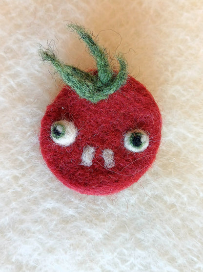 Needle felted wool red tomato brooch with buckteeth on a white knitted fabric background. The brooch has large bulging eyes, two white teeth, and a tuft of fluffy green leaf 'hair'