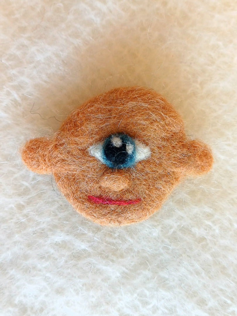 A needle felted wool brooch of a face with dark beige skin, stick-out ears, and one large blue eye. He's smiling calmly. 