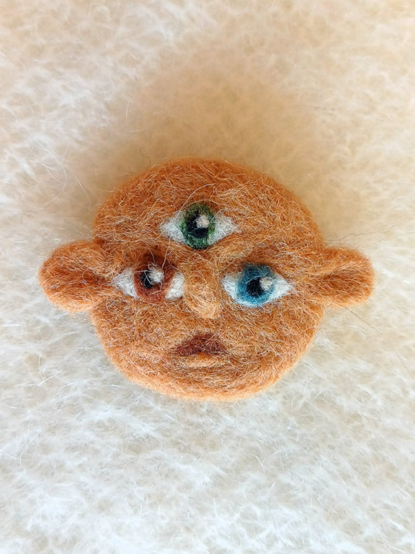Small needle felted face with dark beige skin and 3 eyes; one green, one brown and one blue. His ears stick out either side and he carries a surprised expression.