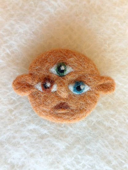 Small needle felted face with dark beige skin and 3 eyes; one green, one brown and one blue. His ears stick out either side and he carries a surprised expression.
