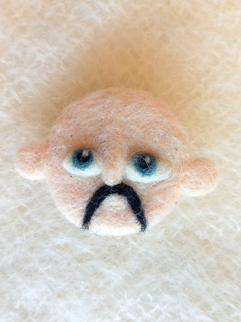 A needle felted wool brooch of a face with pale beige skin, stick-out ears, large blue eyes, and a black handlebar moustache. 