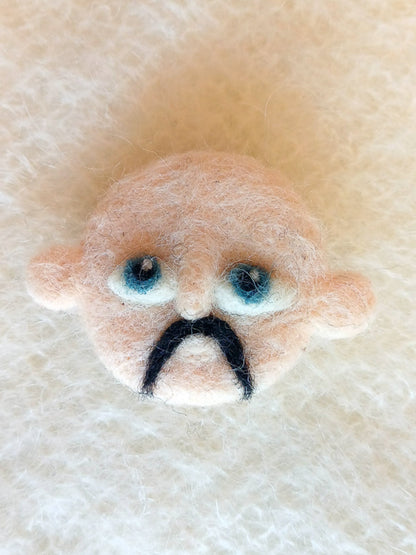 A needle felted wool brooch of a face with pale beige skin, stick-out ears, large blue eyes, and a black handlebar moustache. 
