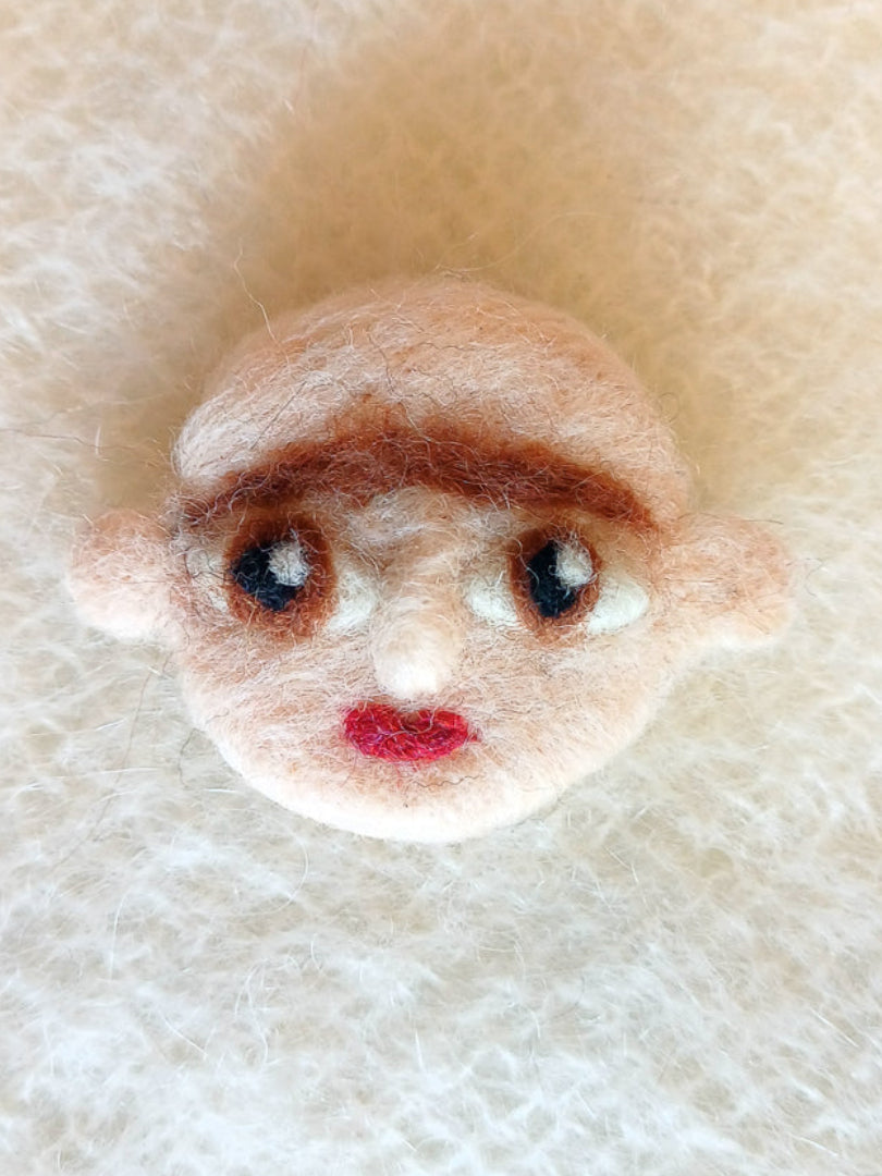 A needle felted wool brooch of a face with pale beige skin, stick-out ears, large brown eyes, red lips and a brown monobrow