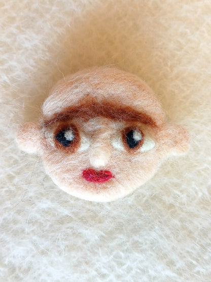 A needle felted wool brooch of a face with pale beige skin, stick-out ears, large brown eyes, red lips and a brown monobrow