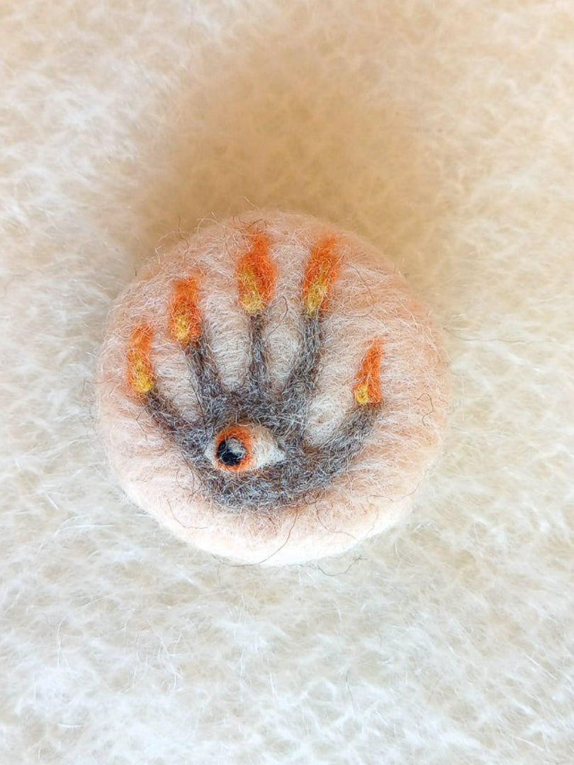 Needle felted cream-white brooch featuring a grey hand of glory with flaming fingers and an orange eye in its palm.