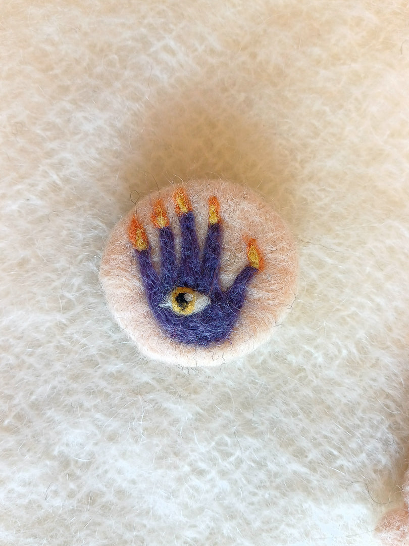 Needle felted cream-white brooch featuring a purple hand of glory with flaming fingers and an orange eye in its palm.