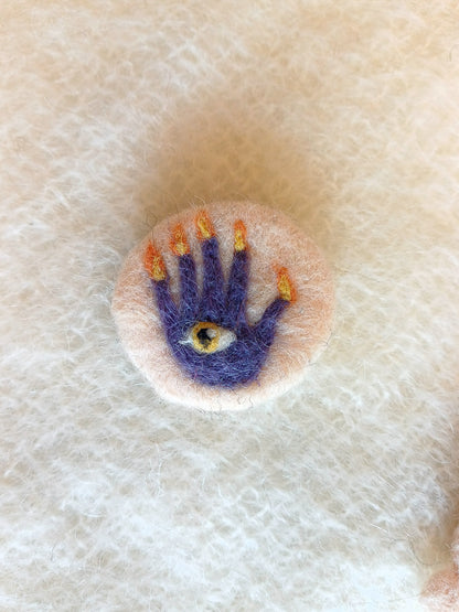 Needle felted cream-white brooch featuring a purple hand of glory with flaming fingers and an orange eye in its palm.