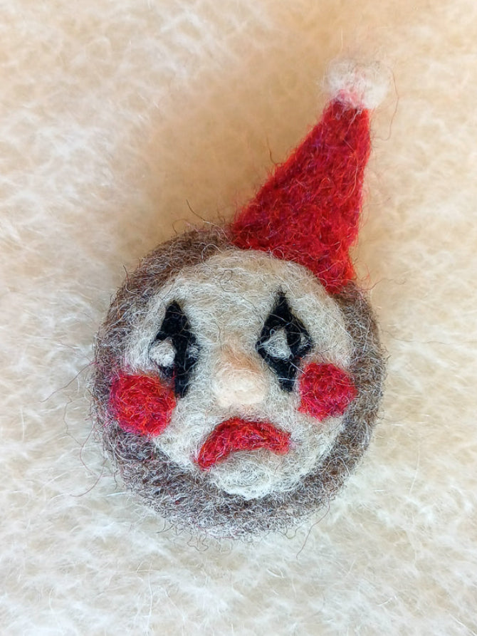 Needle felted clown face brooch with a red hat, red frown and black diamonds on the eyes, on a textured beige background