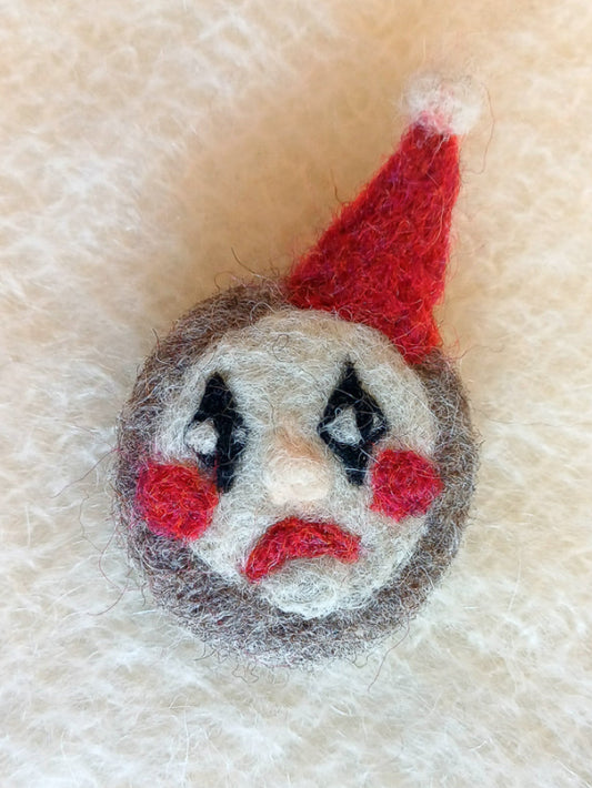 Needle felted clown face brooch with a red hat, red frown and black diamonds on the eyes, on a textured beige background