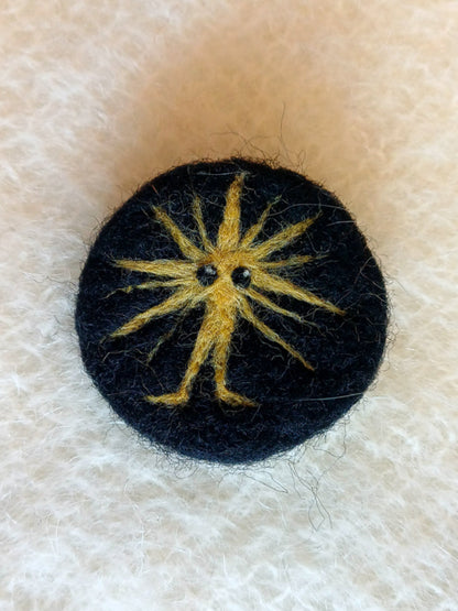 Black needle felted brooch featuring a yellow star burst with feet and two large eyes. 