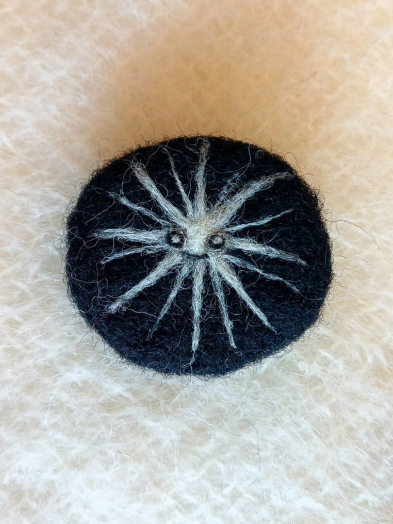 needle felted black round brooch featuring a white star burst with tiny eyes and smile. 