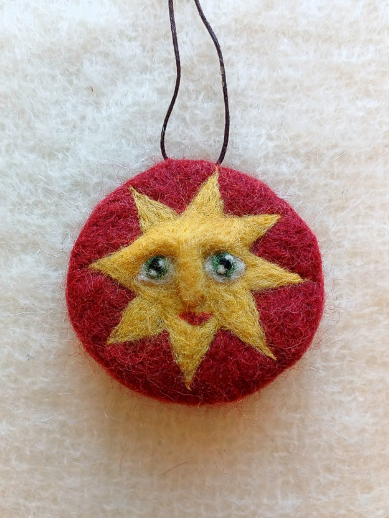 Red needle felted hanging decoration featuring a golden sun face with green eyes. 