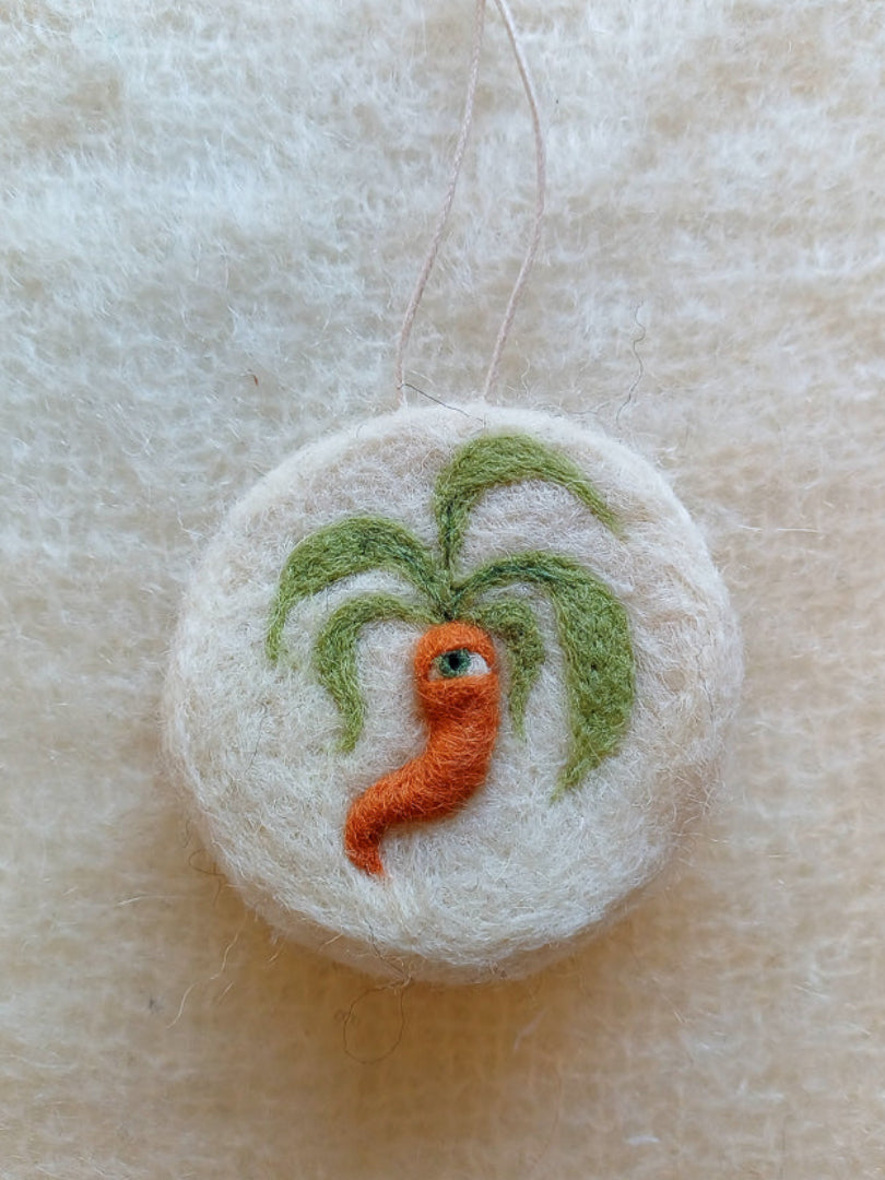 A white circular felted ornament featuring a medieval style carrot with one green eye.