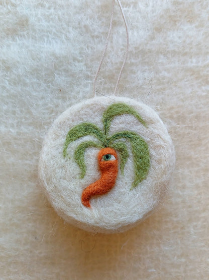 A white circular felted ornament featuring a medieval style carrot with one green eye.