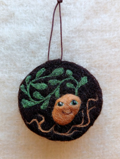 A needle felted brown hanging decoration, featuring an anthropomorphic smiling potato plant, leaves and roots.