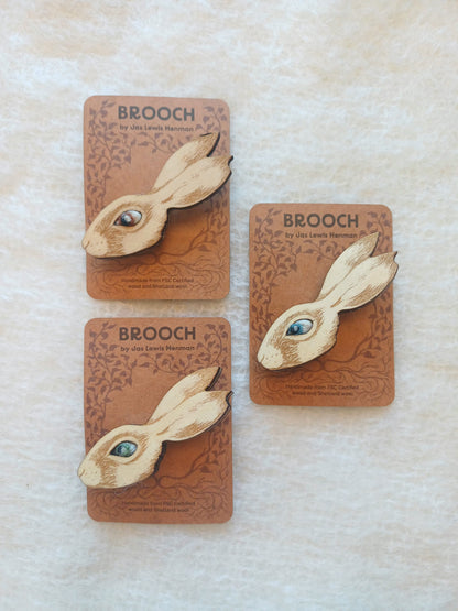 Three wooden rabbit-shaped brooches with different coloured needle felted wool eyes, each mounted on a brown backing card labelled BROOCH, arranged on a light textured surface.