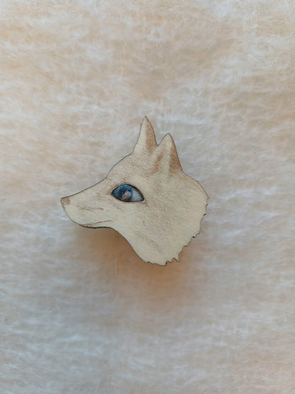 A small wooden fox head pin badge brooch with a blue wool needle felted eye, shown on a soft beige textured background.