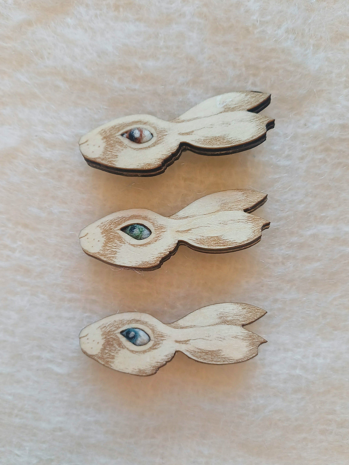 Three small wooden rabbit-shaped items with painted eyes in different colors, arranged vertically on a textured beige surface.