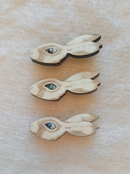 Three small wooden rabbit-shaped items with painted eyes in different colors, arranged vertically on a textured beige surface.