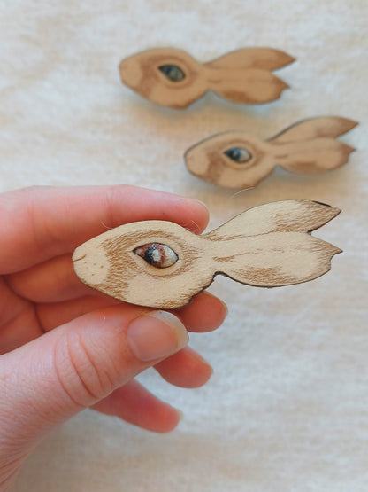 A hand holds a small wooden hare-shaped brooch in the foreground, with two more similar wooden hare brooches blurred in the background on a light fabric surface.