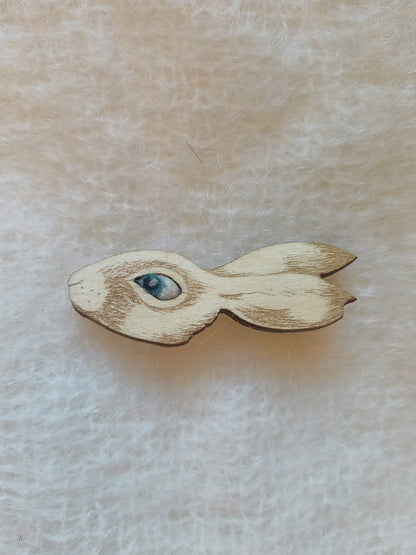A small wooden rabbit-shaped brooch with a detailed blue and white eye, displayed on a soft, textured cream-colored fabric background.