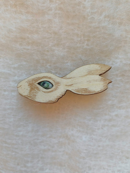 A small wooden rabbit-shaped brooch with a detailed laser-cut face and a green-blue stone for an eye, resting on a soft beige textured fabric background.