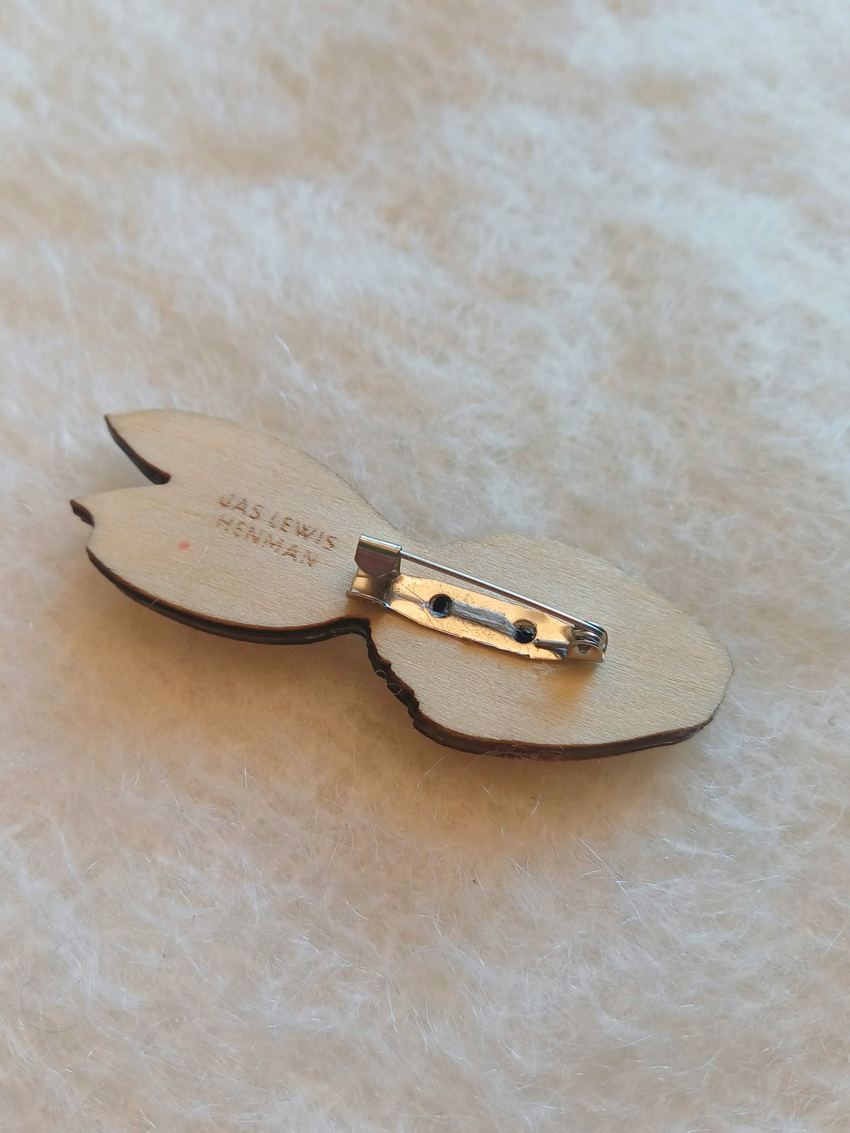 A small wooden pin badge with sewn on clasp/fastener, showing the back side with the brand name 'JAS LEWIS HENMAN' etched in it, resting on a soft white textured fabric background.