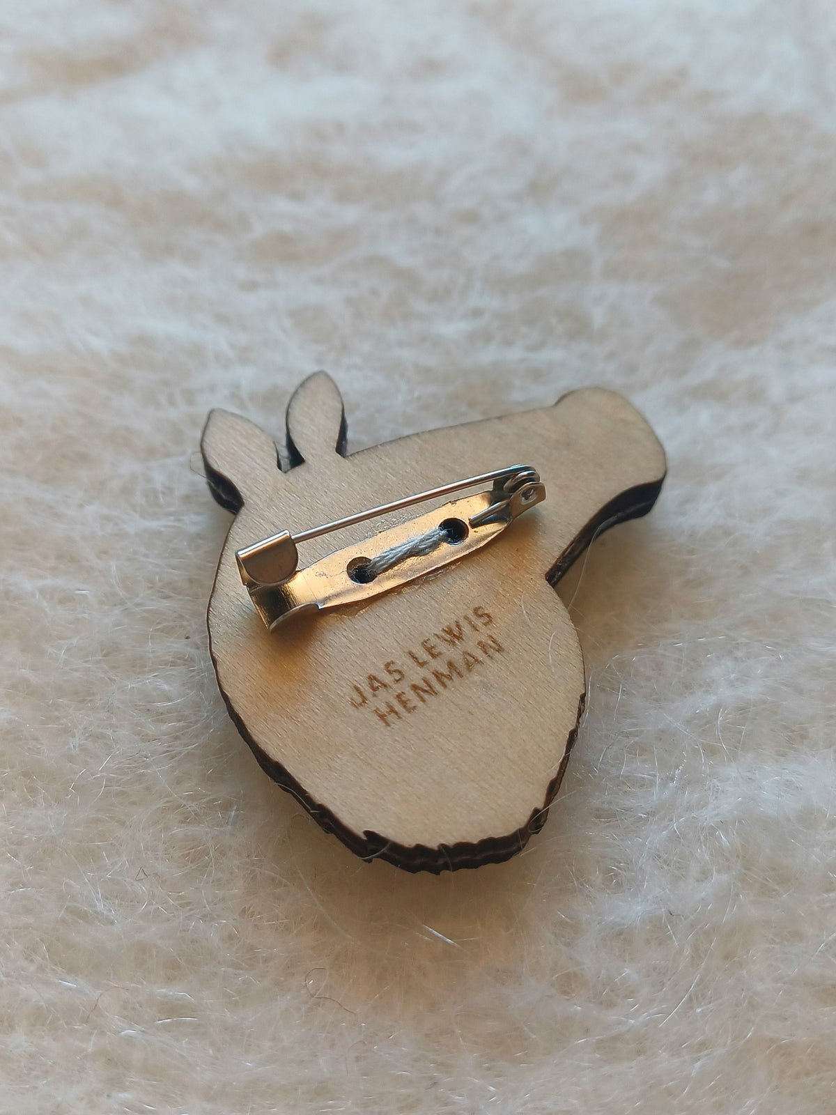 The back of a wooden boar-shaped badge with a safety pin c-clasp, featuring the engraved text 'JAS LEWIS HENMAN' on a soft, textured cream background.