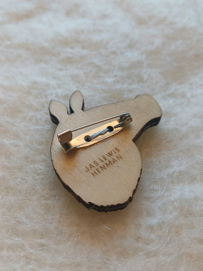 The back of a wooden boar-shaped badge with a safety pin c-clasp, featuring the engraved text 'JAS LEWIS HENMAN' on a soft, textured cream background.