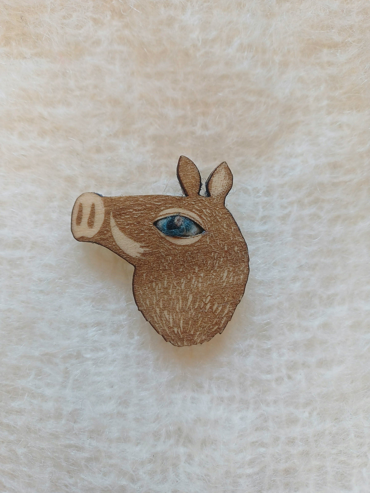A small wooden boar head pin badge brooch with a blue wool eye, shown on a textured off-white fabric background.