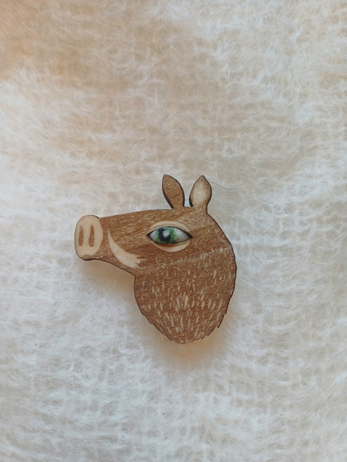 A small wooden boar head brooch with a detailed green woollen eye, displayed on a textured cream-colored fabric background.