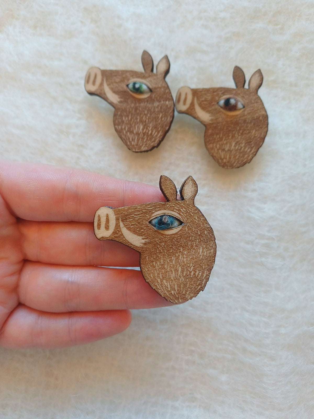 Three small wooden boar head brooches with needle felted wool eyes are shown on a soft cream fabric background, with one brooch being held in a person's hand for scale.