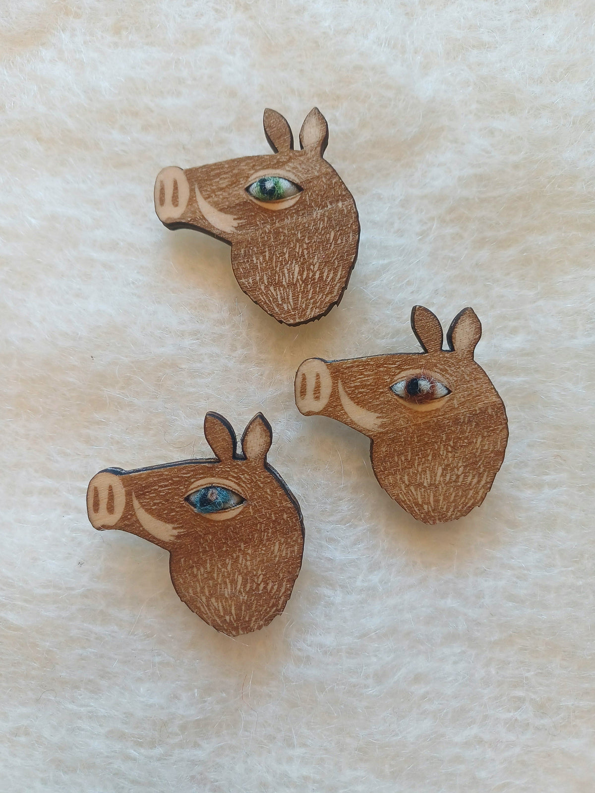 Three wooden boar head brooches with colourful wool eyes, displayed on a textured cream fabric background.