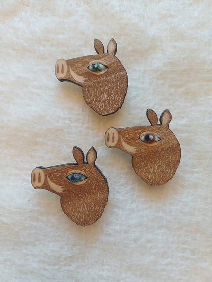 Three wooden boar head brooches with colourful wool eyes, displayed on a textured cream fabric background.