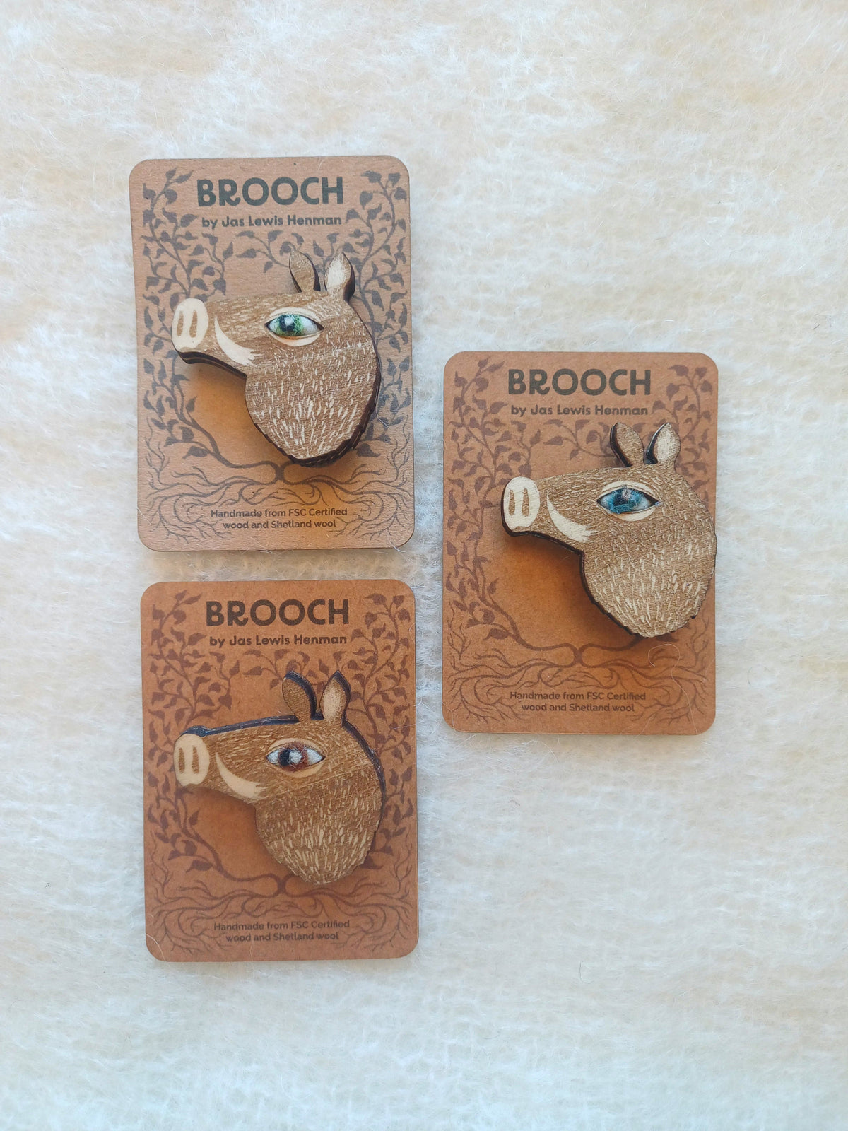 Three wooden boar brooches with colorful wool needle felted eyes, mounted on decorative brown display cards labelled BROOCH by Jas Lewis Henman, arranged on a light textured fabric background.