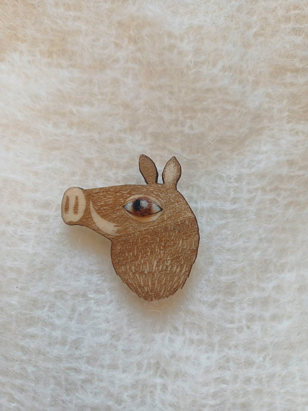 A small wooden pig-shaped pin badge or brooch with a detailed wool eye and engraved texture, resting on a soft, textured off-white fabric background.