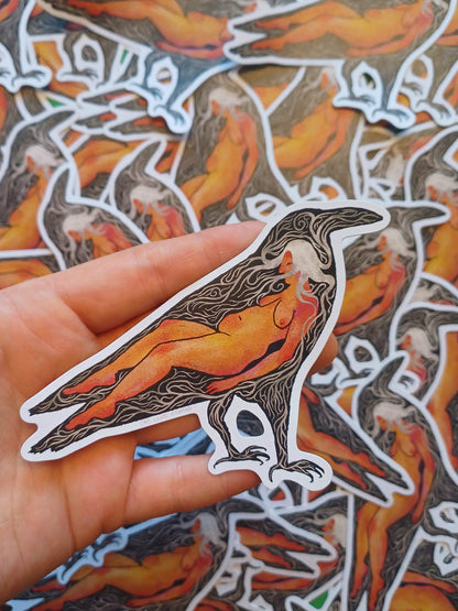 A hand holds a die-cut sticker featuring a stylized raven with a nude female figure inside, rendered in orange, black, and white. Multiple identical stickers are scattered in the background.