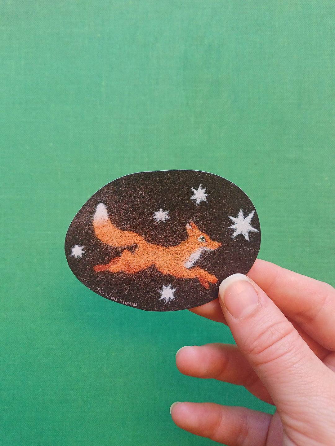 A hand holding a dark brown oval sticker featuring an orange fox running among white stars, set against a solid green background.