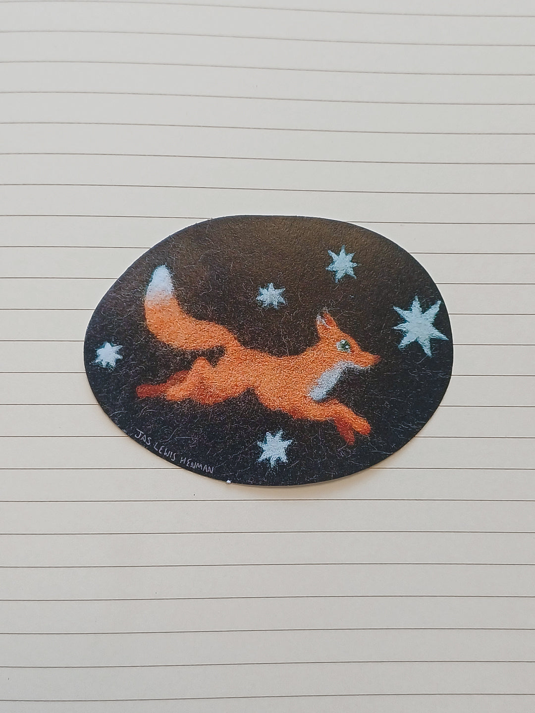 An oval sticker featuring a running orange fox on a dark brown background with light blue-white stars, placed on a lined notebook page.