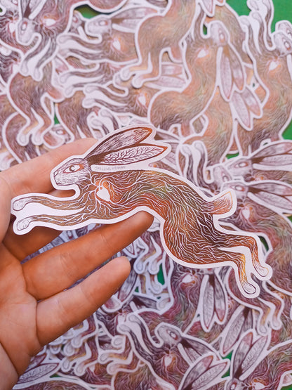 A hand holds a die-cut sticker or decal of a stylized rabbit in warm brown, orange, and cream tones, with many similar rabbit cutouts scattered in the background.
