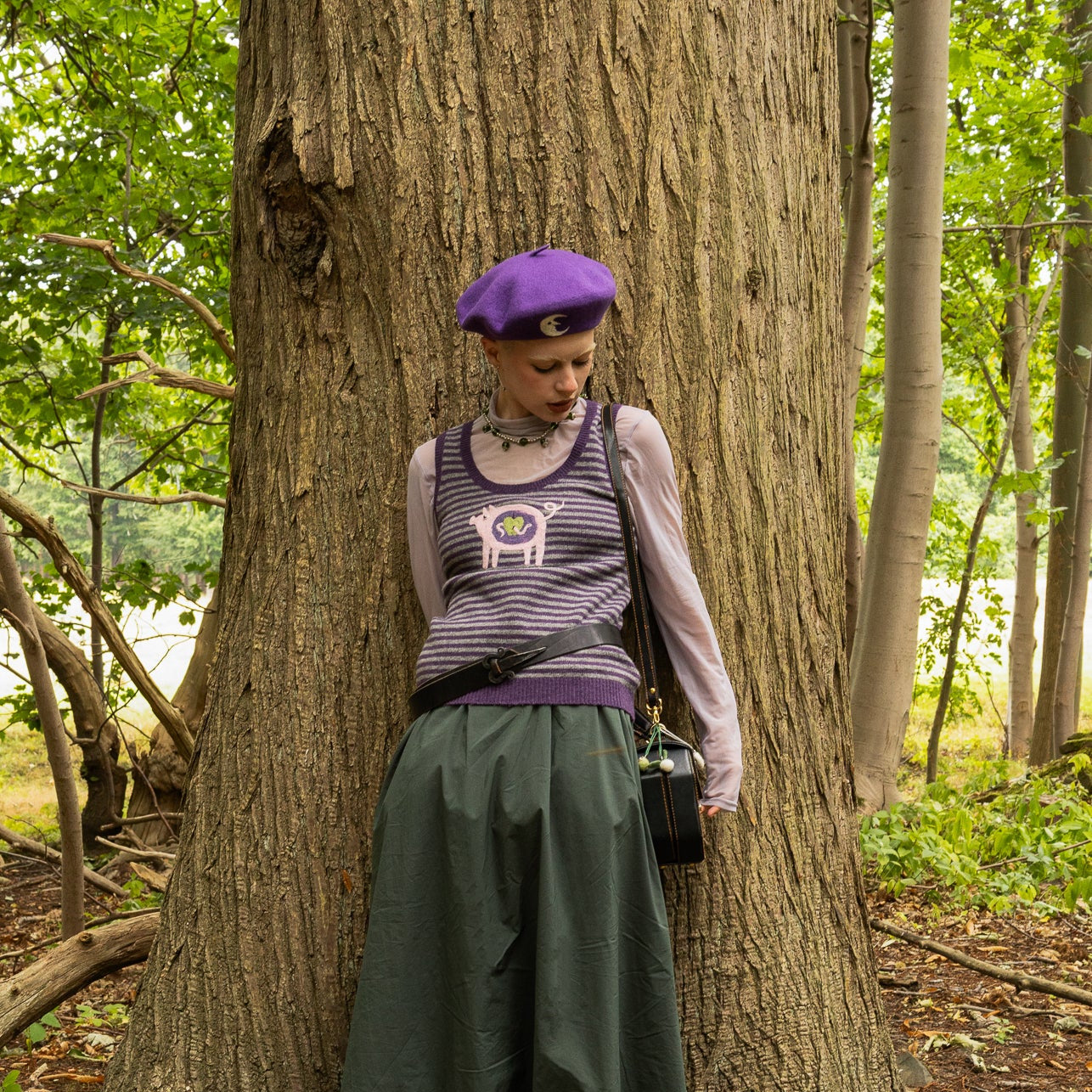Person wearing the purple crescent moon beret, upcycled piggy vest and green skirt standing next to a tree in a forest