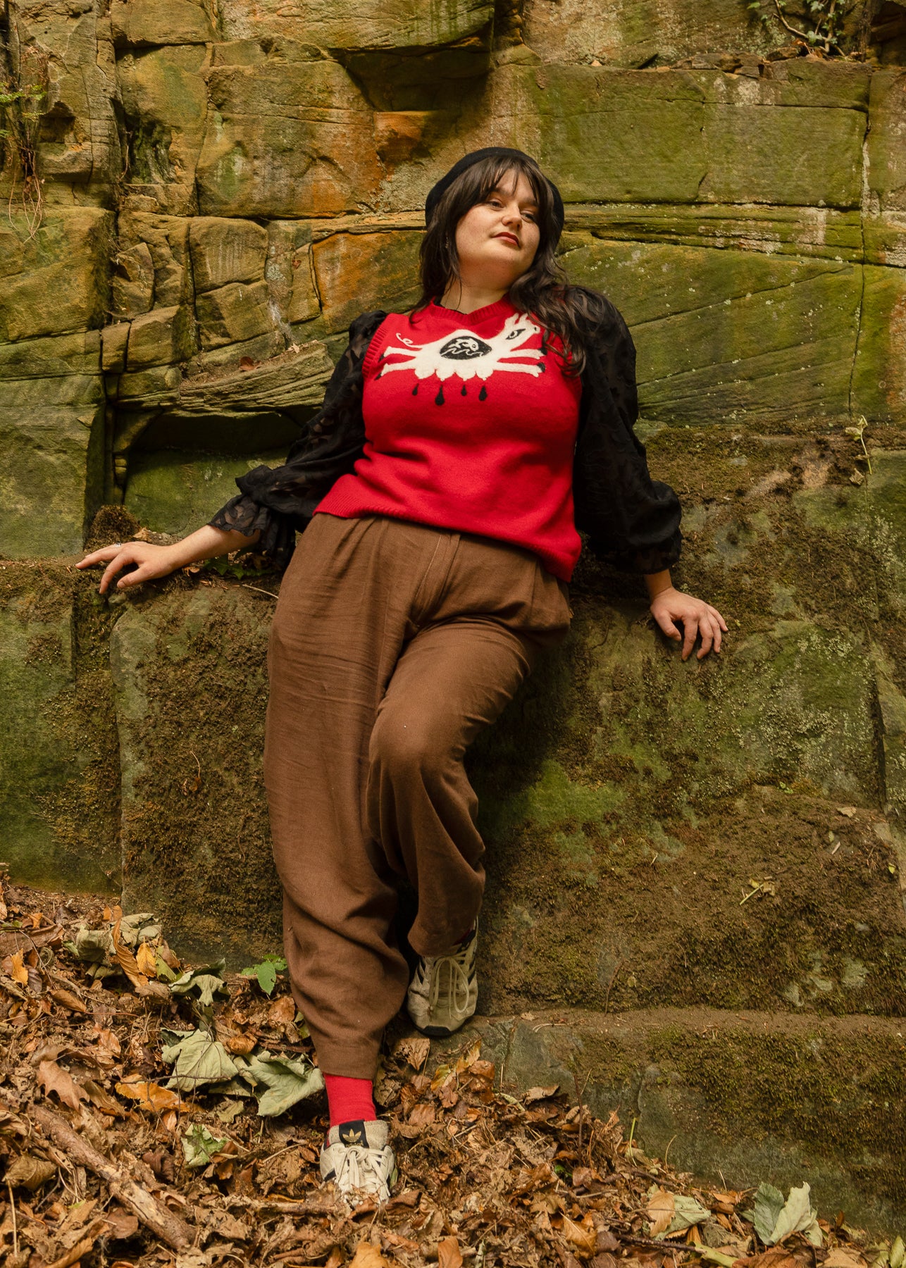 Jas Lewis Henman wearing a red knitted vest upcycled with a white needle felted pig, and brown linen trousers, leaning against a stone wall.