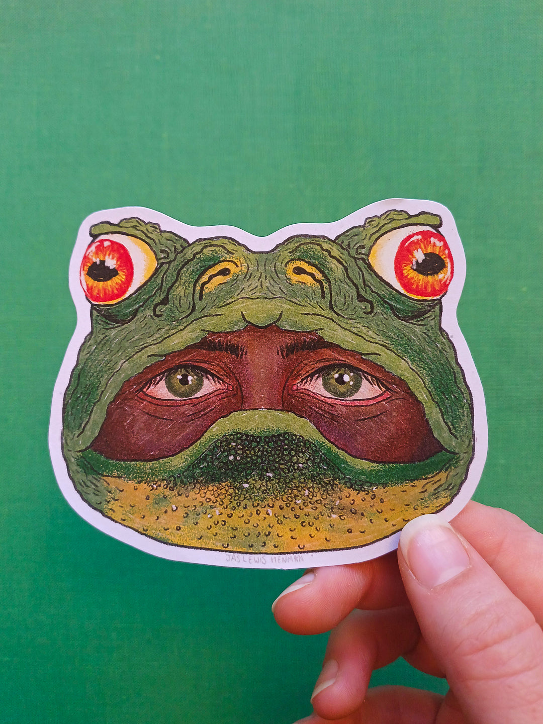 A hand holds a die-cut sticker shaped like a frog head with surreal human eyes in it's mouth, shown against a solid green background.