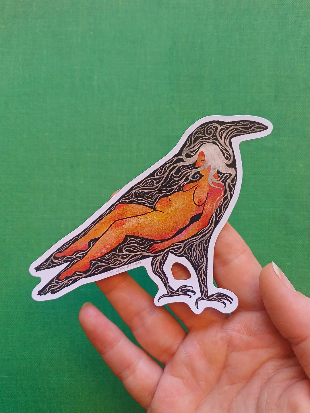 A die-cut sticker featuring a stylized raven with an orange and yellow nude figure inside, held in a person's hand against a solid green background.