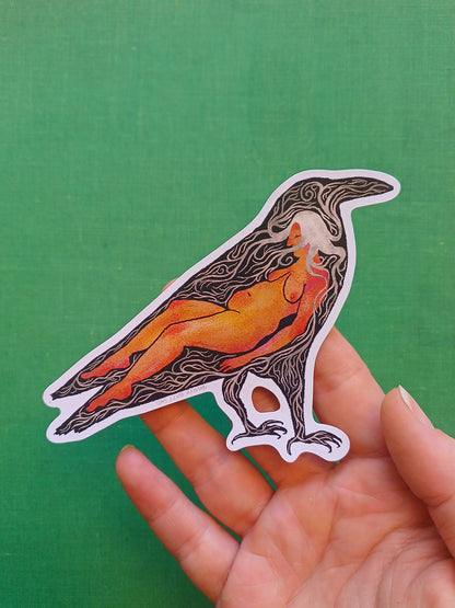 A die-cut sticker featuring a stylized raven with an orange and yellow nude figure inside, held in a person's hand against a solid green background.